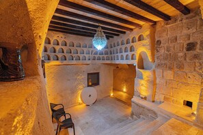 A stone-walled room with a chandelier, arched niches, a fireplace, and a wooden ceiling.