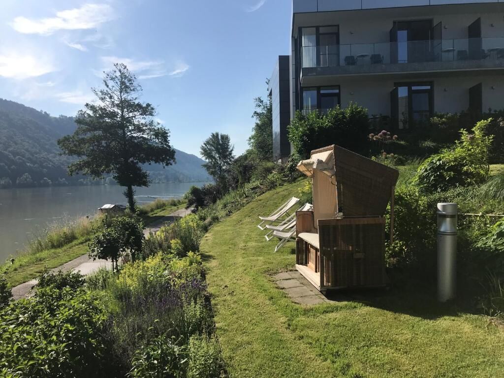 Un edificio moderno con balcón, un área verde con sillones de descanso y vistas a un lago.