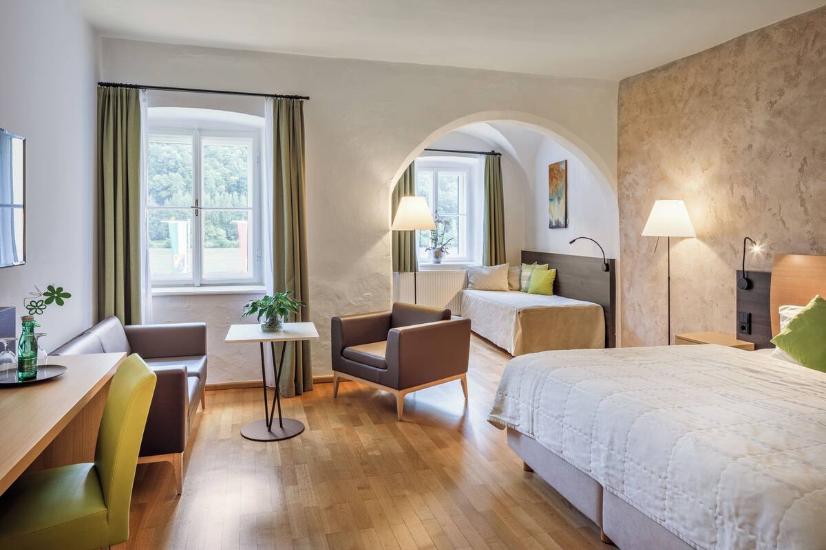 Ein modernes Hotelzimmer mit Holzboden, einem Bett, einer Couch, einem Sessel, einem Tisch und einem Fenster mit Vorhängen.