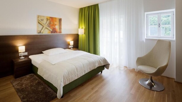 A hotel room with a bed, a chair, a nightstand, a lamp, a painting, and a window with curtains. - Hotel Wesenufer (Wesenufer)