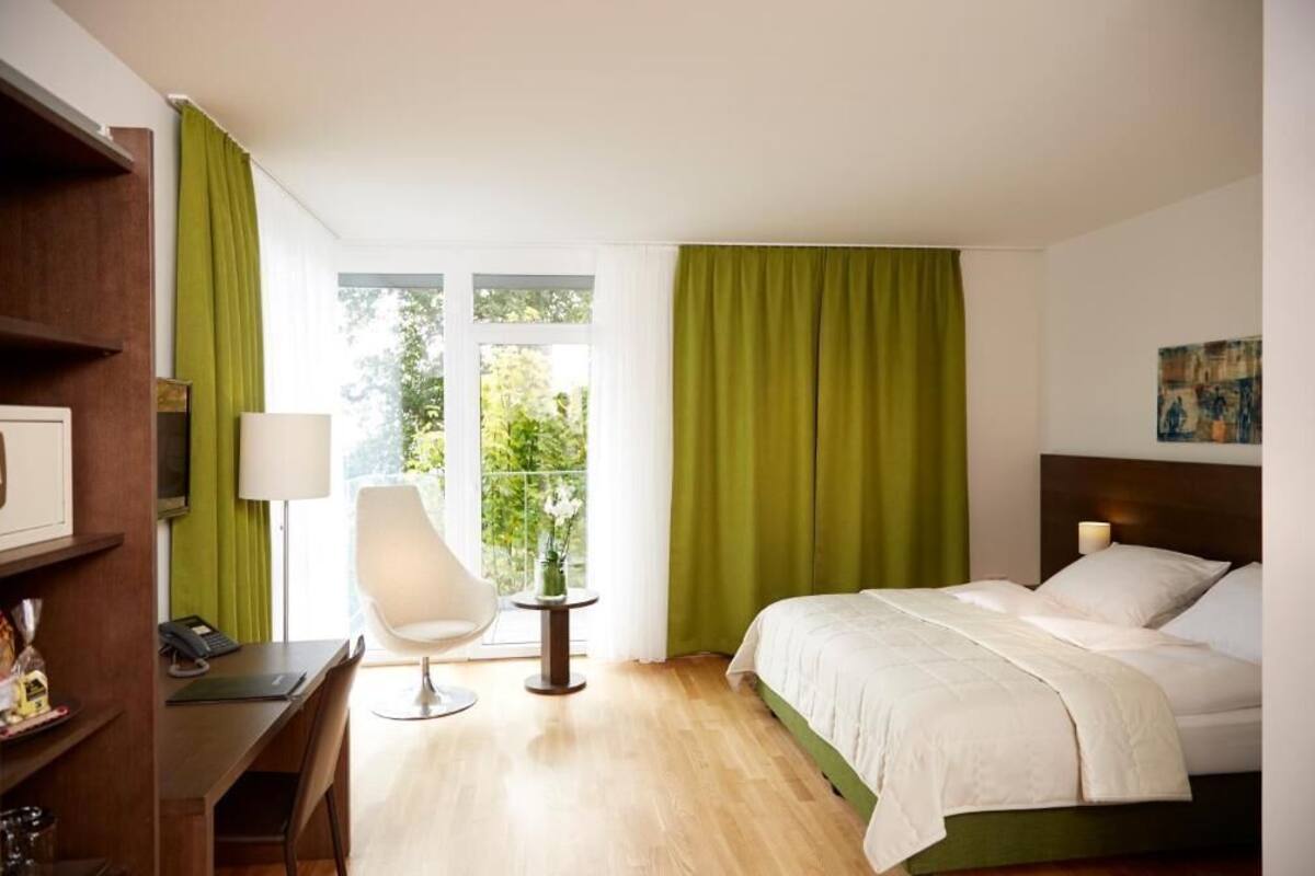 Ein Hotelzimmer mit Bett, Schreibtisch, Stuhl, Lampe und einem Fenster mit Vorhängen.