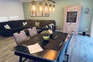 A modern living room with a sectional sofa, a dining table with placemats, and a built-in wooden cabinet. - Holiday home in Pewsum with swimming pool (Pewsum)