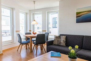 A modern living room with a sofa, dining table, chairs, and a pendant light. - Apartment building Sandperle (Norderney)
