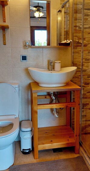 Ein Badezimmer mit Toilette, einem Holzwaschtisch mit Waschbecken, einer Duschkabine aus Glas und einer kleinen Mülltonne.