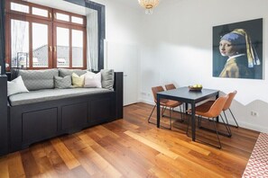 Ein Zimmer mit einem dunkelgrauen Sofa, einem Esstisch mit Stühlen, einer Kristallleuchtre und einem großen Gemälde an der Wand.