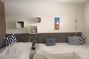 A hotel room with two beds, a striped headboard, a wall-mounted shelf with books, a painting of a lighthouse, and a star-shaped decoration. - Holiday apartment with 2 bedrooms (Borkum, Deutschland)
