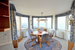 Ein Essbereich mit einem runden Tisch und sechs Stühlen, großen Fenstern mit Vorhängen und Blick aufs Meer.