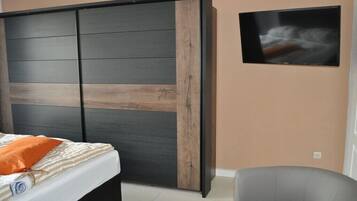 Ein Schlafzimmer mit einem Bett, einem grauen Sessel, einem an der Wand befestigten Fernseher und einer Schiebetür mit Holzoptik.