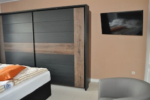 Ein Schlafzimmer mit einem Bett, einem grauen Sessel, einem an der Wand befestigten Fernseher und einer Schiebetür mit Holzoptik.