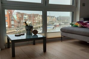Een kamer met uitzicht op een jachthaven, een glazen tafel met een fles en onderzetter, een grijze bank en een kleurrijk kussen.