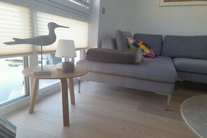 Ein modernes Wohnzimmer mit einem grauen Sofa, einem kleinen runden Holztisch mit einer Lampe und einer dekorativen Vogel-Skulptur.