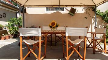 Eine überdachte Terrasse mit Holztisch und vier Stühlen, umgeben von Kübelpflanzen.
