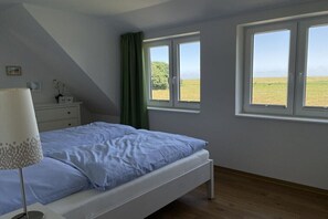 A bedroom with a bed, a bedside table, a lamp, two windows, and a view of a field. - Sea view in the Emma holiday home in Arngast (Varel-Dangast)
