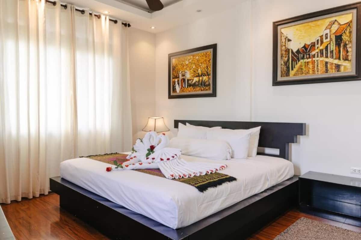 Una cama bien hecha con sábanas blancas y una toalla decorativa, una lámpara de noche y un cuadro en la pared.