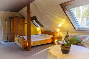 A bedroom with a wooden bed, a nightstand, a bedside lamp, a potted plant, and a window with a view of trees. - "Studio" in the "White House Varwig" (Nebel\/Süddorf)
