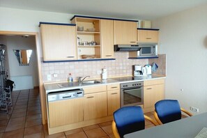Eine kleine Küche mit Holzschränken, einer Mikrowelle, einem Herd und einem Spülbecken. Zwei blaue Stühle stehen an einem Tisch.
