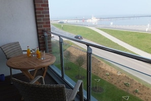 Ein Balkon mit Blick auf eine Straße und eine Brücke, ein Tisch mit Getränken, Korbstühle und ein Glasgeländer.