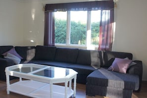 Ein Wohnzimmer mit einem dunkelgrauen Sofa, einem Glastisch und einem Fenster mit Vorhängen.