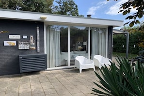 Ein modernes Haus mit schwarzem Anstrich, einer Glastür, die in einen Wohnbereich führt, und einer Terrasse mit weißen Stühlen.
