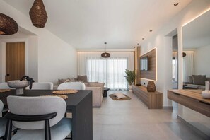 Un salon moderne comprenant un canapé, une table à manger et des chaises, un téléviseur fixé au mur et une grande fenêtre ornée de rideaux légers.