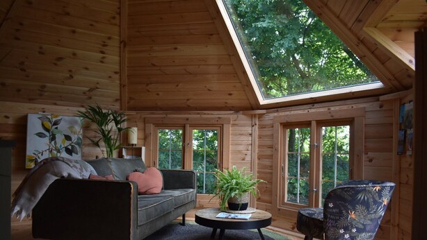 A cozy cabin interior with a wooden ceiling, a sofa, a chair, a small table, and a large window. - Finnish kota with a private wood-fired barrel sauna (Slootdorp)