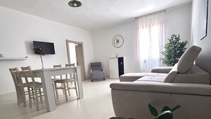 Un salon comprenant un canapé, une table à manger avec des chaises, un téléviseur fixé au mur et une plante.