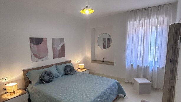 Une chambre à coucher avec un lit, des lampes de chevet, un miroir rond, une fenêtre avec des rideaux légers et une table en bois.