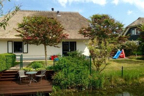 Ein Haus mit einem Strohdach, einer Terrasse mit Stühlen, einem kleinen Teich und einem Spielplatz mit einer Rutsche.