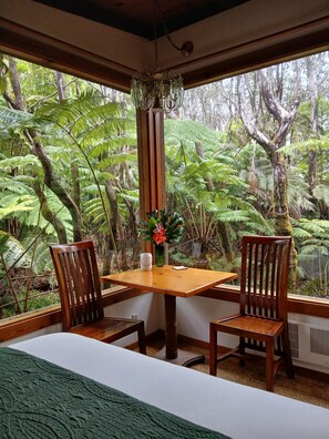 A room with a wooden table and chairs, a hanging light fixture, and a view of a dense forest through large windows.