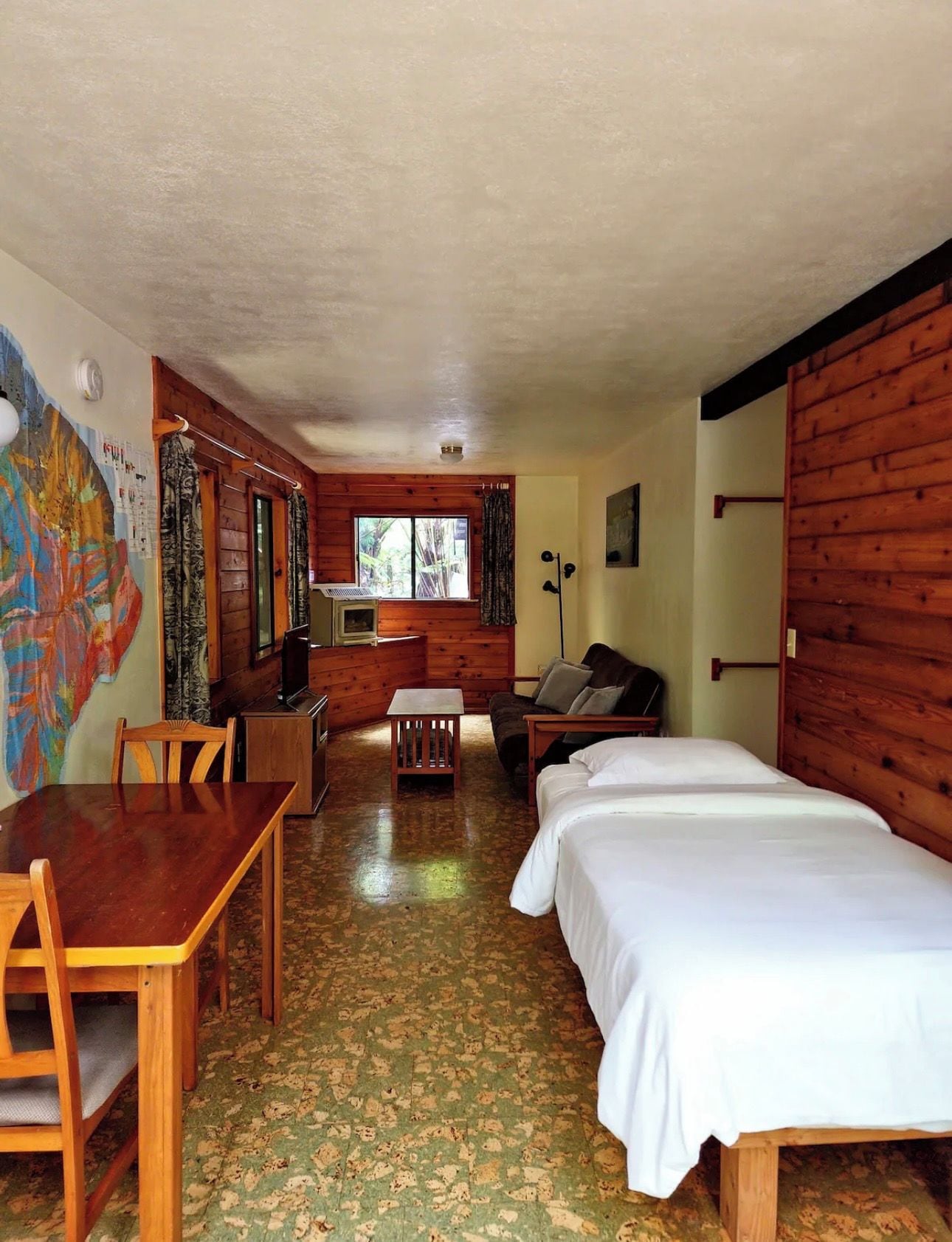 A room with a wooden floor, a bed, a desk, a chair, a table, a window, and a wall with a colorful mural.