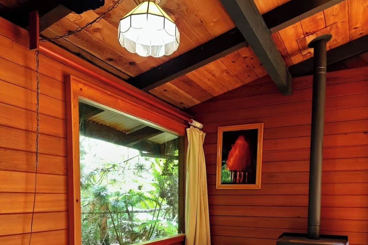 A wooden cabin room with a bed, a window with curtains, a painting, and a wood-burning stove.