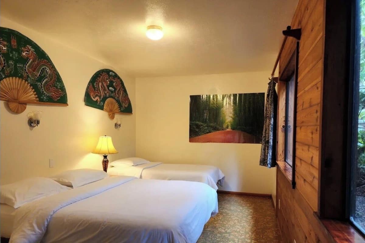 A room with two beds, a lamp, a painting, and a window with curtains.