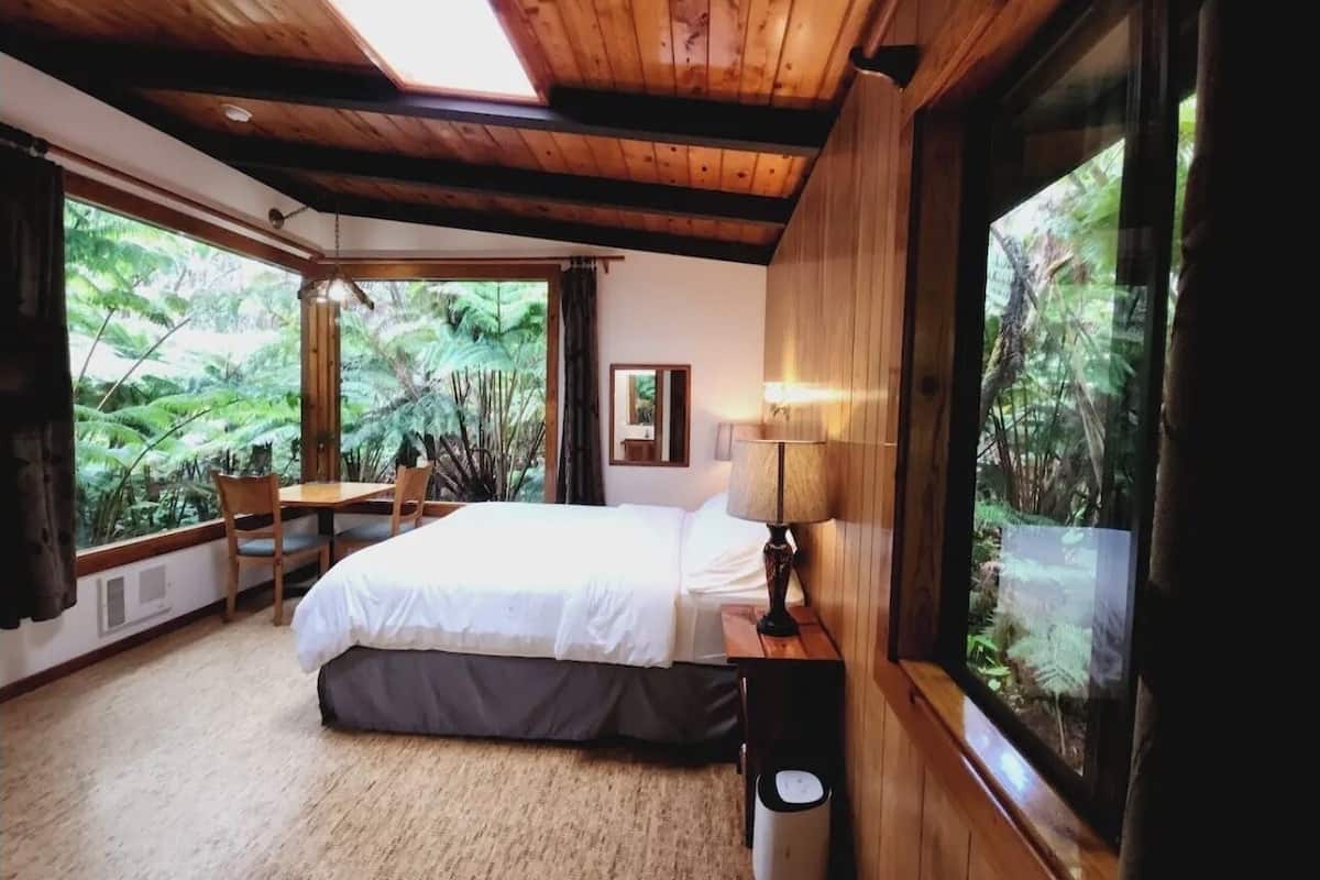 A bedroom with a bed, bedside table, lamp, and a window with a view of greenery.