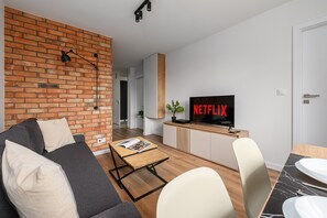 Un salon moderne avec un mur en briques, un canapé, une table basse, un téléviseur affichant le logo « NETFLIX », et un coin repas agrémenté d’une plante.