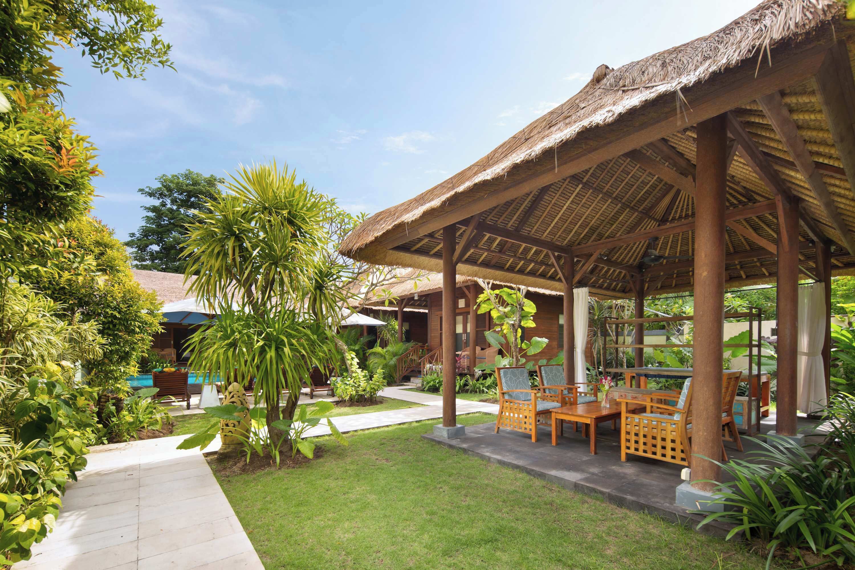 A covered outdoor seating area with wooden furniture, surrounded by lush greenery and a clear sky.