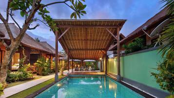 A pool area with a covered seating area, a thatched-roof building, and lush greenery.