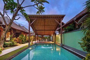 A pool area with a covered seating area, a thatched-roof building, and lush greenery. - Gecko Bungalow (Nusa Lembongan)