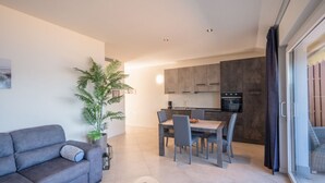Ein modernes Wohnzimmer mit einer Couch, einem Esstisch mit Stühlen, einer Küchenzeile mit Schränken und einem großen Fenster mit Vorhängen.