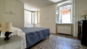 Ein Schlafzimmer mit Bett, Heizkörper, einem Fenster mit Vorhängen, Schreibtisch mit Lampe und einem Schminktisch.