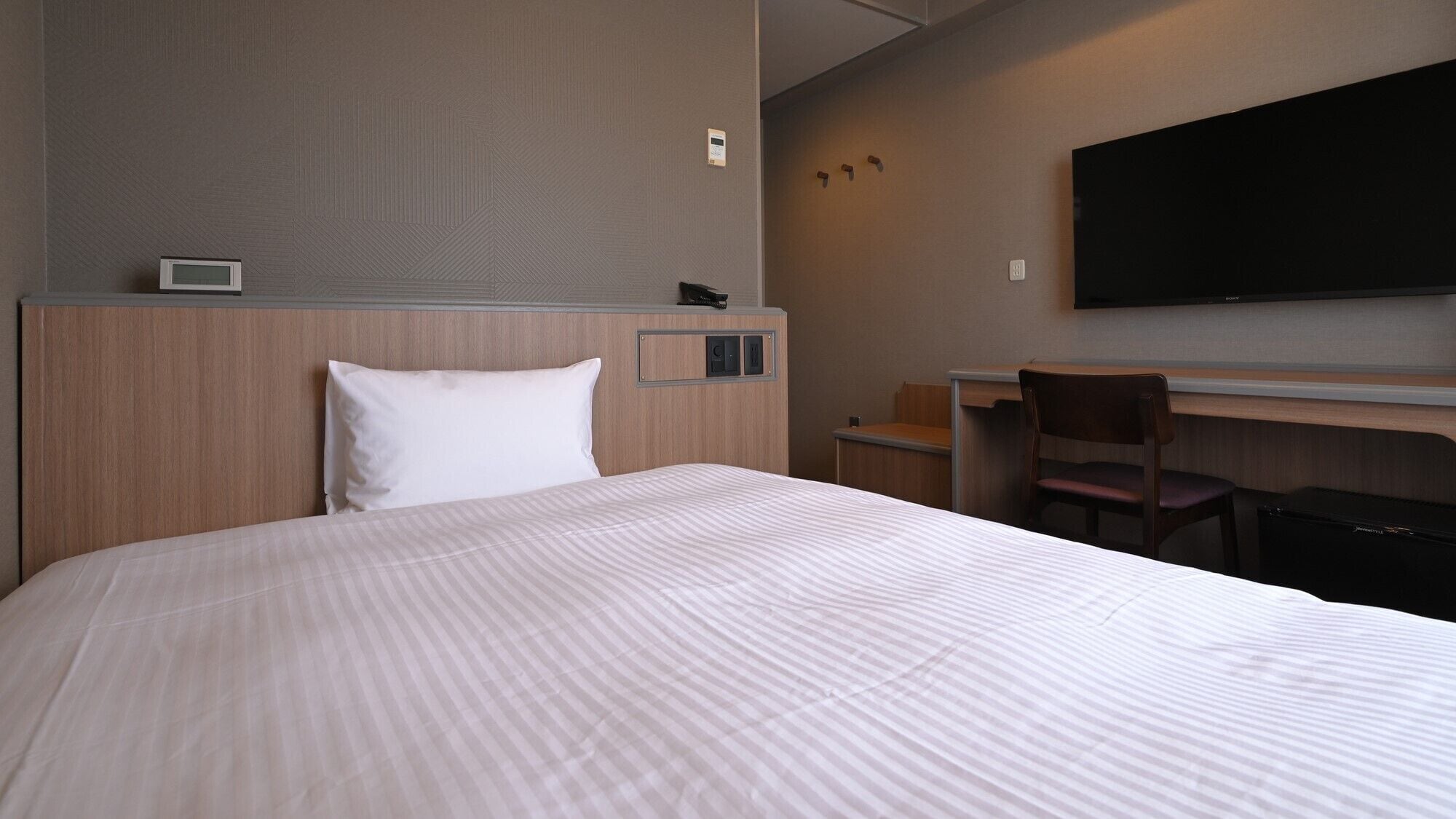 A hotel room with a bed, a desk, a chair, a TV, and a wall-mounted control panel.