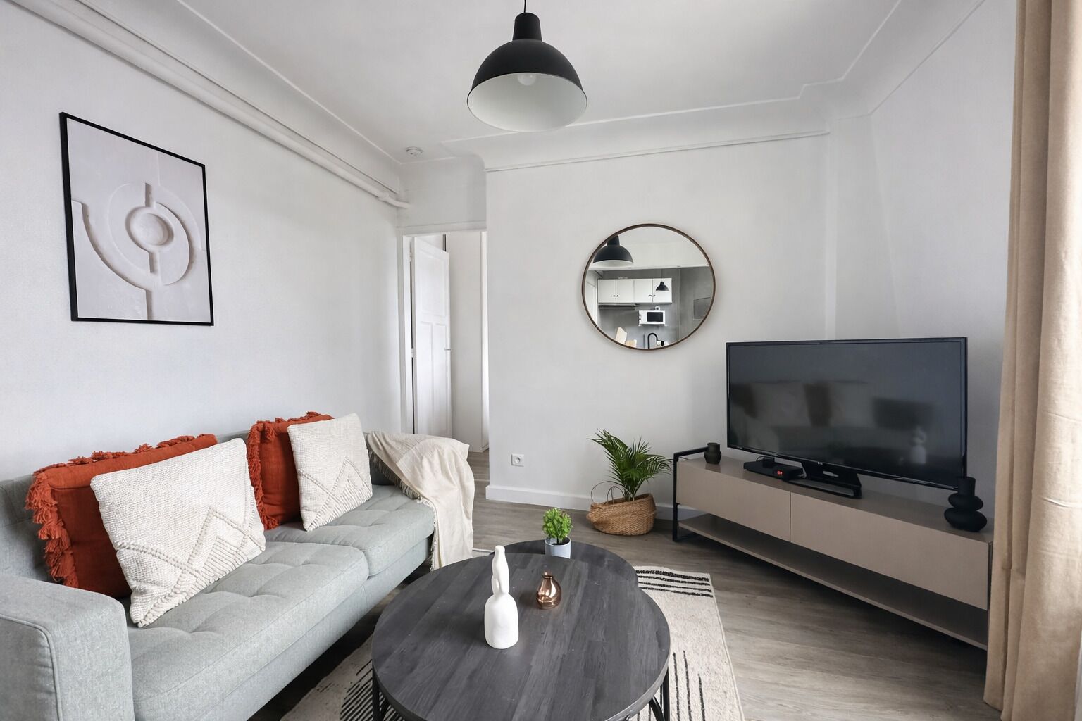 A modern living room with a grey sofa, a round coffee table, a flat-screen TV, a mirror, and a pendant light.