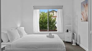 A white bedroom with a bed, a window with curtains, a bedside table with a lamp, and a wall-mounted shelf. - Spacious 3-Bed Home in Payneham South (Payneham South)
