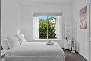 Quarto branco com cama, janela com cortinas, mesa de cabeceira com abajur e prateleira na parede.