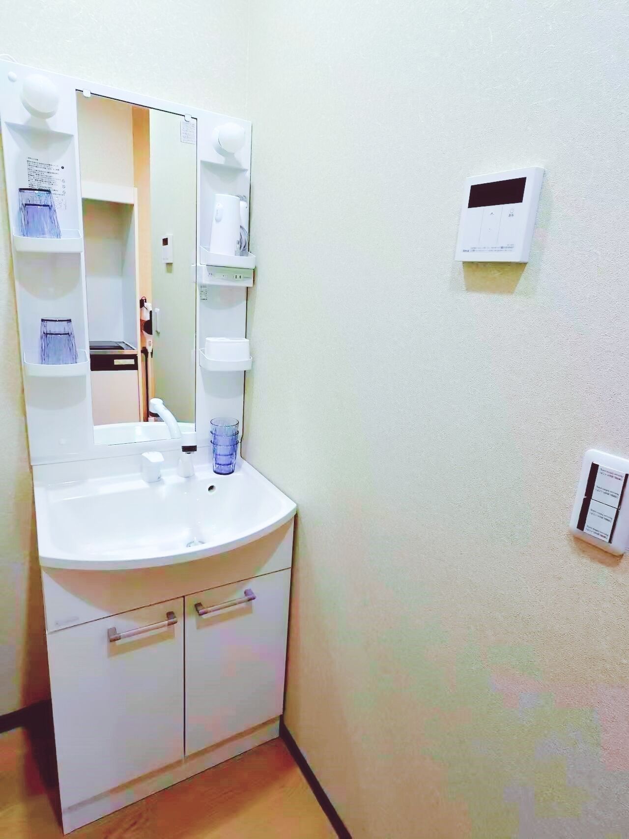 A bathroom with a white sink, mirror, and two towel racks. There is a wall-mounted thermostat and a light switch.