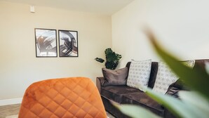 Een woonkamer met een donkerbruine bank, twee abstracte kunstwerken in lijst, een grote groene plant en een oranje fauteuil.
