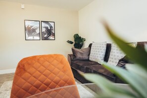 Ein Wohnzimmer mit einem dunkelbraunen Sofa, zwei gerahmten abstrakten Kunstwerken, einer großen Grünpflanze und einem orangefarbenen Sessel.