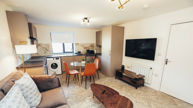 Een moderne woonkamer met een bank, salontafel en een eethoek met stoelen. In de achtergrond is een keuken met een fornuis, spoelbak en koelkast zichtbaar.