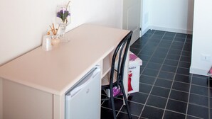 Una habitación con un escritorio blanco, una silla, un televisor montado en la pared, una puerta y suelo de baldosas.