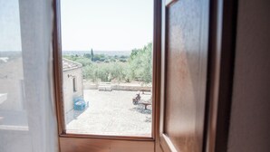Vista a través de una ventana de un patio con suelo de piedra, bancos y un edificio al fondo.