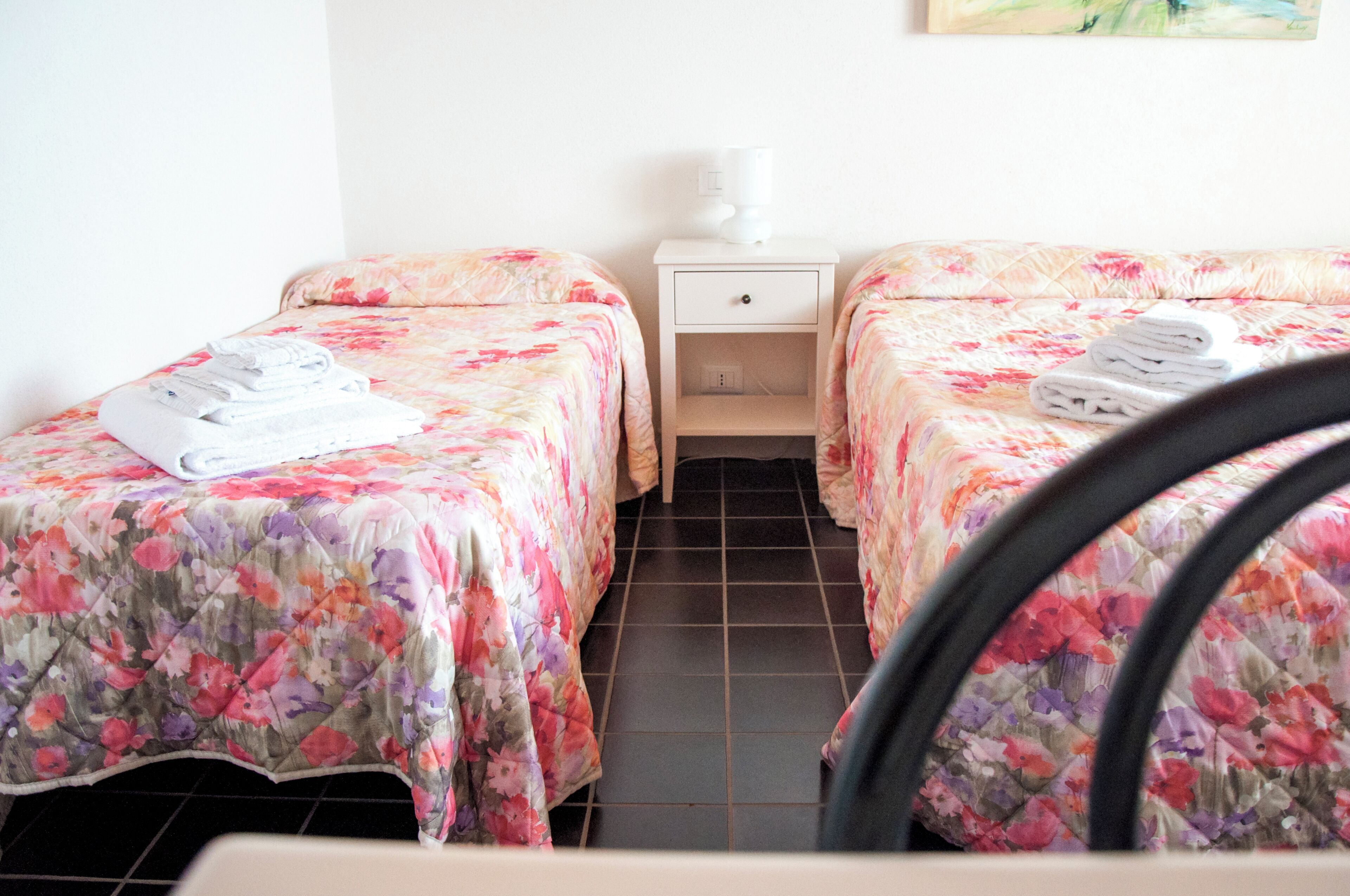 Een hotelkamer met twee bedden, elk met een bloemenprint deken en een opgevouwen handdoek op het bed. Er staat een witte nachtkastje met een lamp tussen de bedden.
